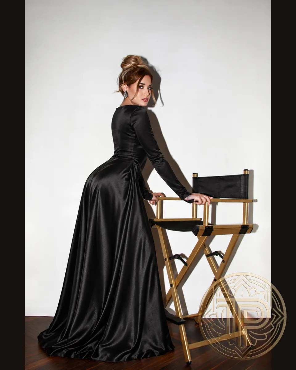 Maya in a long black satin dress, looking over her shoulder while standing next to a gold director's chair